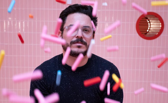 A man with glasses and a beard looks forward as colorful pink, blue, and yellow cylinders float around him.