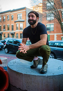 A bearded man in a black t-shirt and olive pants sits on a concrete structure in a city.