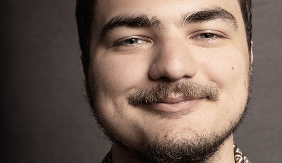 Close-up portrait of a man with dark eyebrows, a mustache, and a light beard smiling.
