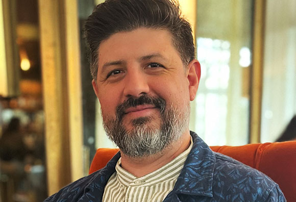 A man with dark hair and a grey-flecked beard wearing a blue patterned shirt smiles.