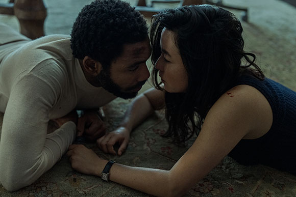 A man in a white long-sleeve shirt and a woman in a dark tank top lie on a patterned floor, looking intensely at each other.