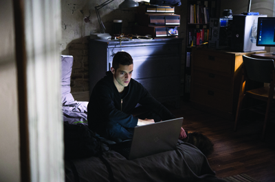 A man in a dark hoodie sits on the floor using a laptop in a dimly lit room with a desk and bookshelf.