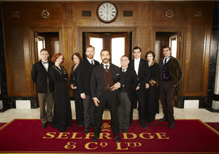 A group of nine people in early 20th-century formal clothing standing on a red carpet in a grand hallway.