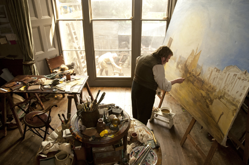 An artist in a studio painting a large landscape canvas on an easel.