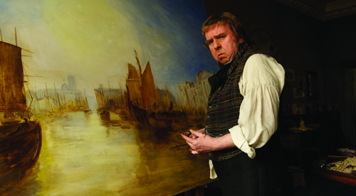 A man in a vest and white shirt stands beside a large painting of a coastal scene with ships.