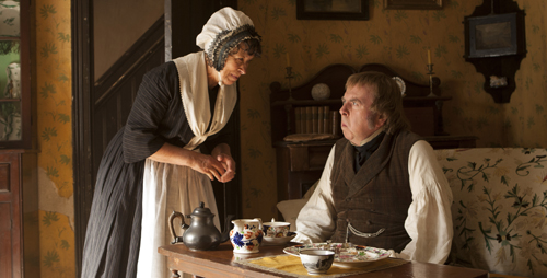 A woman in a white cap and apron leans over a table to serve tea to a man sitting in a dimly lit room.