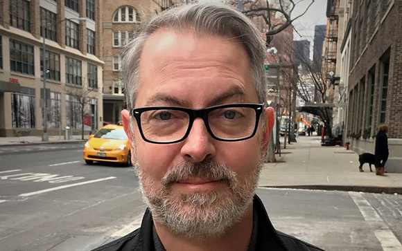 A man with graying hair, a beard, and black-rimmed glasses looks toward the camera on a city street.