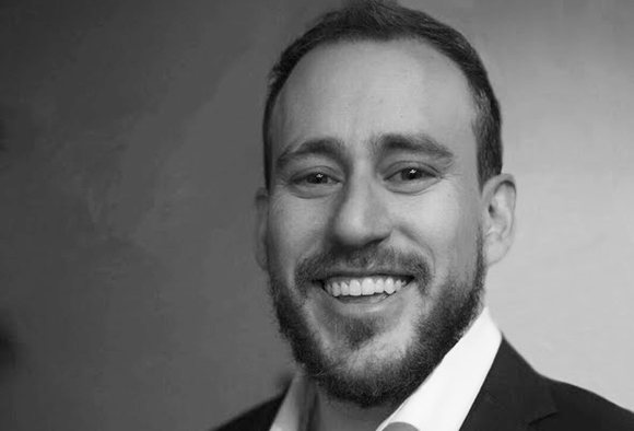 Black and white headshot of a smiling man with a beard and short hair wearing a collared shirt.