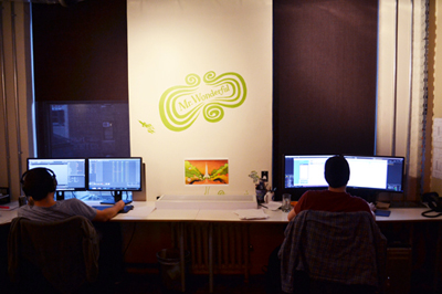 Two people sit at desks with multiple monitors in a dimly lit office with a large logo on the wall.