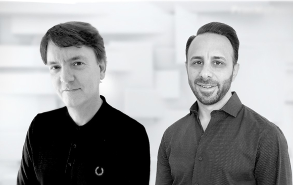 Black and white portrait of two men standing side by side against a light, textured background.