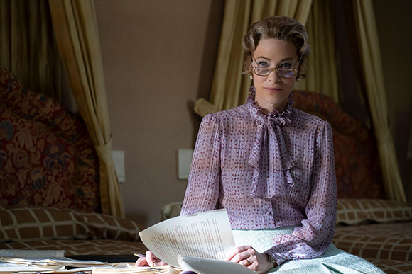 A woman with wavy hair and glasses wearing a purple patterned blouse sits before a canopy bed.