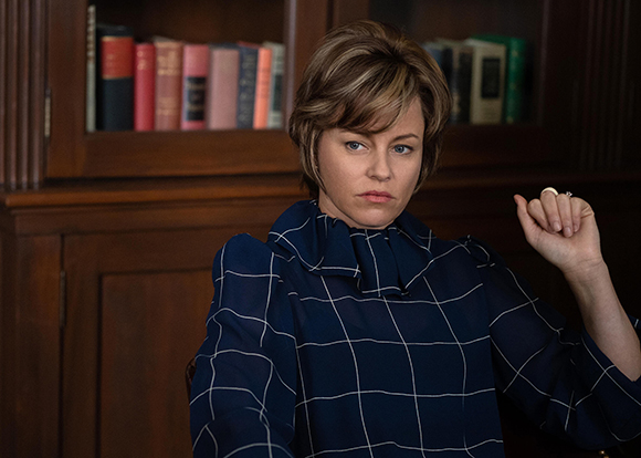 A woman with short brown hair wearing a navy blue grid-patterned blouse sits before a wooden bookshelf.