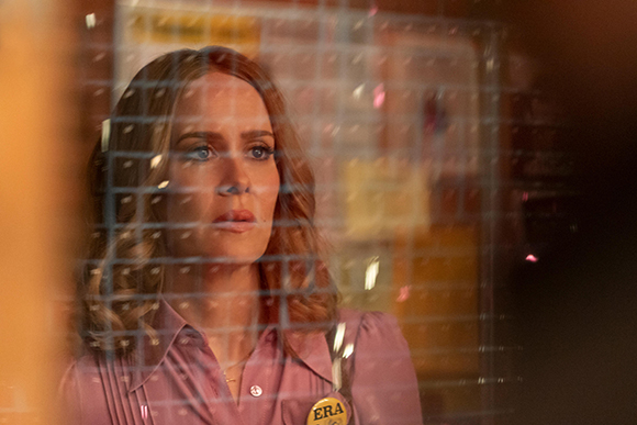 A woman with wavy reddish-brown hair looks through a wire mesh window with a concerned expression.