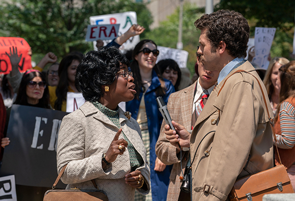 A woman in a white jacket speaks to a man holding a microphone amidst a crowd holding ERA protest signs.