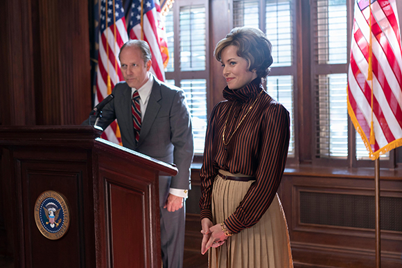 A man in a grey suit speaks at a wooden podium next to a woman in a striped turtleneck and tan skirt.