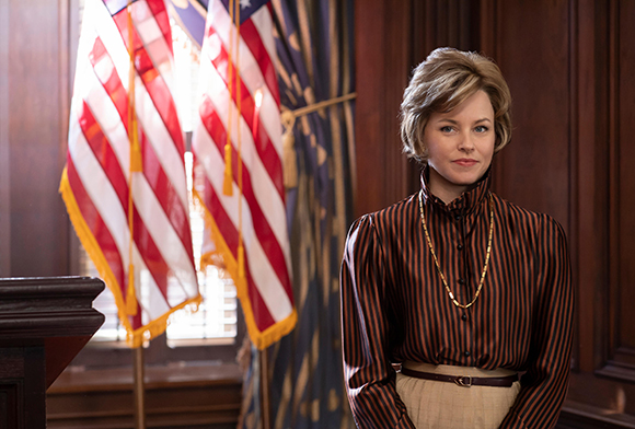 A woman with short blonde hair wearing a striped blouse stands before two American flags in a wood-paneled room.