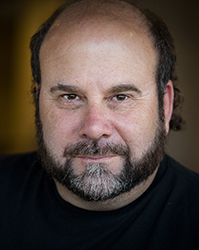 Close-up portrait of a man with dark hair, a beard, and a mustache looking toward the camera.