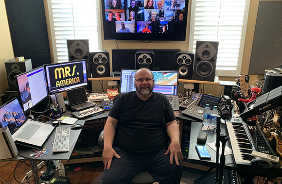A man sits at a desk filled with computer monitors, keyboards, and audio equipment in a studio.
