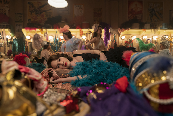A woman in a black dress lies on a bed surrounded by teal feathers and colorful theatrical costumes.