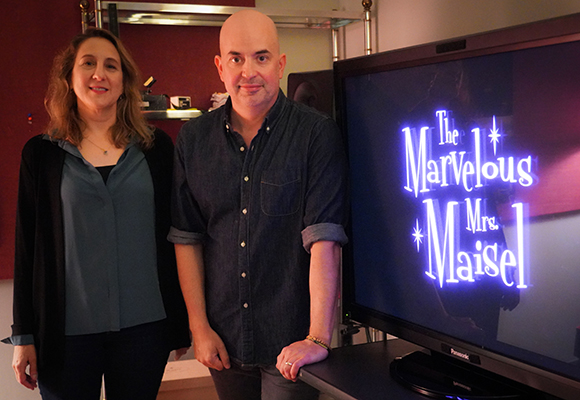 A man and a woman stand beside a television displaying The Marvelous Mrs. Maisel logo.