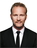 A man with light hair and a mustache wearing a black suit, white shirt, and black tie against a white background.