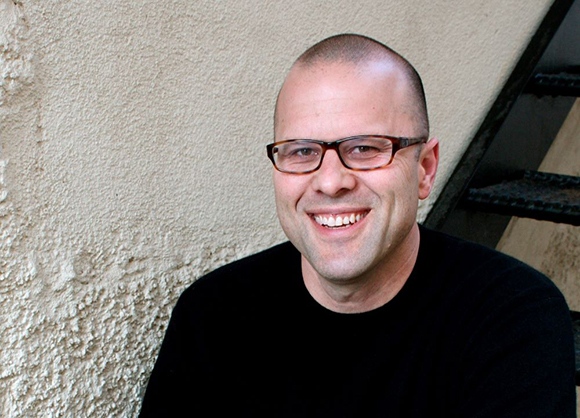 A smiling man with short hair and glasses wearing a black shirt against a textured wall.