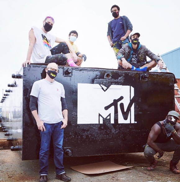 Six people wearing face masks pose around and on top of a large black metal container with an MTV logo.