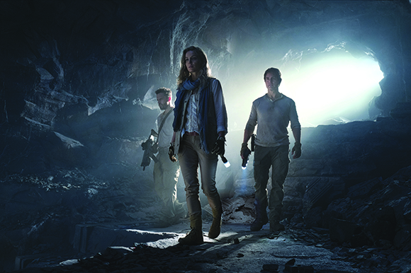 Three people walk through a dark, rocky cave toward a bright opening in the distance.