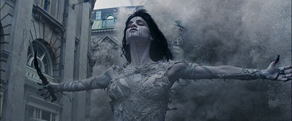 A woman with dark hair and face markings stands with arms outstretched in a cloud of smoke.