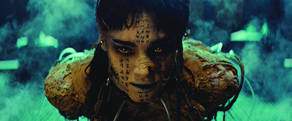 A person with dark hair and glowing yellow eyes, their face covered in dark markings, amidst teal smoke.