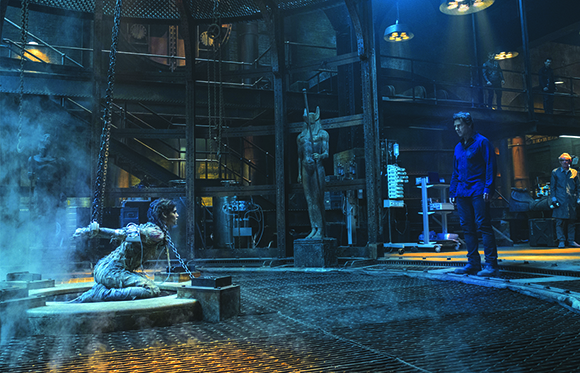 A man in a blue shirt stands in a dark, metallic industrial room looking toward a person in a pit.