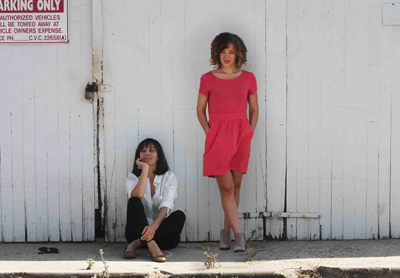 A woman in a red dress stands while another woman in a white shirt sits on the ground against a white wall.