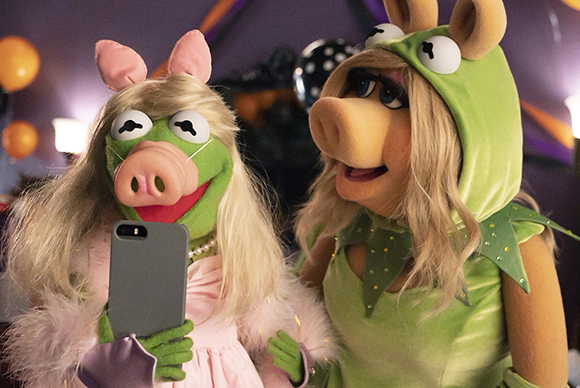 Two green puppet characters in costumes pose for a selfie with a smartphone.