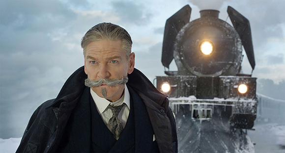 A man with a grey mustache and goatee in period clothing stands before a steaming train in the snow.