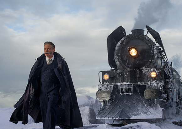 A man in a dark coat walks past a large, snow-covered steam locomotive under a cloudy sky.