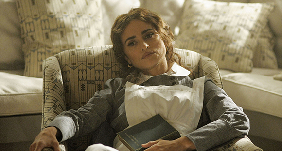 A woman with wavy brown hair sits in a patterned armchair wearing a grey and white dress.