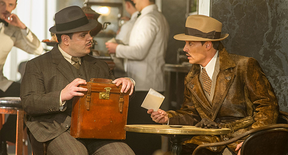 Two men in 1930s suits and hats sit at a cafe table, one holding a brown leather briefcase.