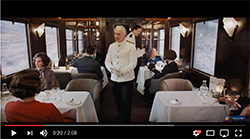 A waiter in a white jacket walks through a dining car with passengers seated at tables.