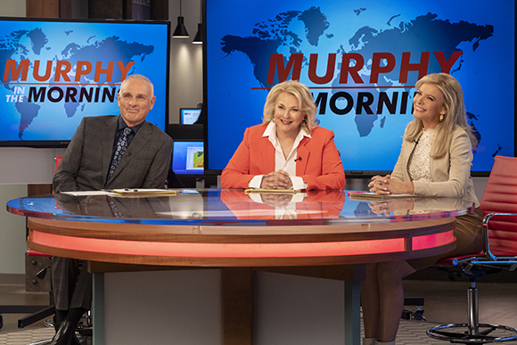 Three news anchors sit behind a curved desk in front of a blue world map background.