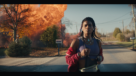 A woman in a purple tank top runs down a road as a large orange explosion erupts behind her.