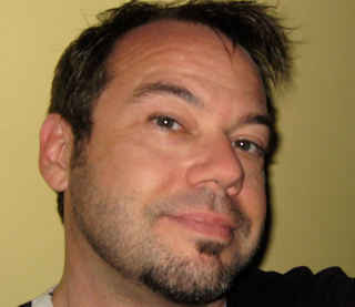 A man with short dark hair and light facial stubble looking toward the camera against a yellow background.