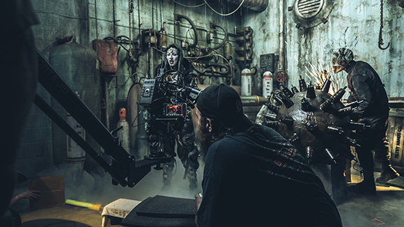 A camera operator films an actress in costume on a dark, industrial movie set with sparks flying.