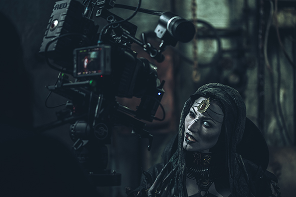 A person in dark gothic makeup and hooded attire looks toward a large film camera on a set.