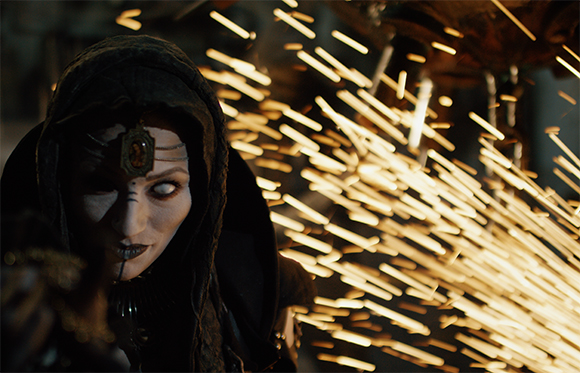 A pale, hooded figure with white eyes looks forward amidst a spray of bright, glowing sparks.