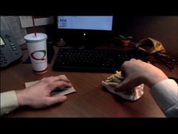 A person's hands eating a sandwich at a desk next to a computer monitor, keyboard, and a large drink.