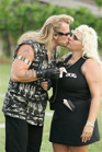 A blonde man in a patterned vest and a blonde woman in a black dress kiss outdoors.