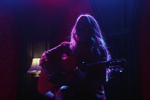 A person playing an acoustic guitar in a dark room lit by pink and blue lights.