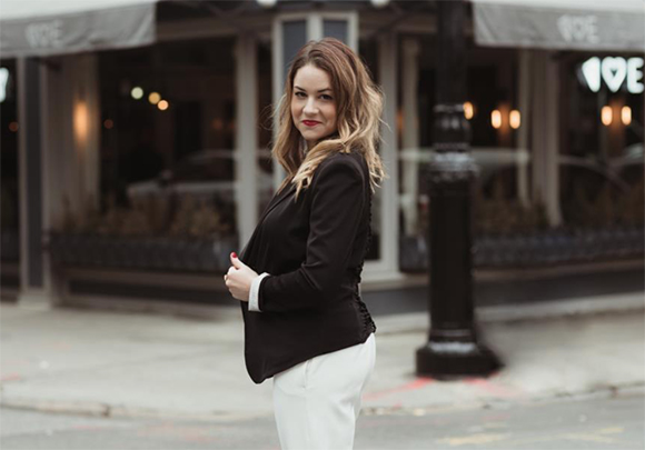 A woman with wavy blonde hair wearing a black blazer and white trousers, looking at the camera on a city street.