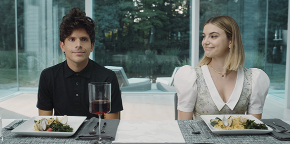 A man in a black polo and a woman in a white blouse sit at a table with plates of pasta and red wine.