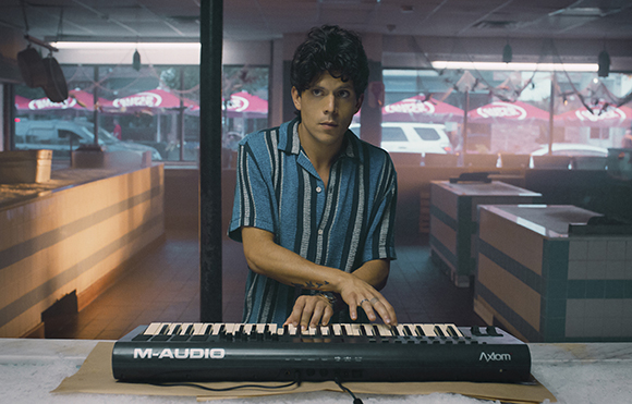A man in a striped shirt plays a black M-Audio Axiom keyboard in a room with large windows.